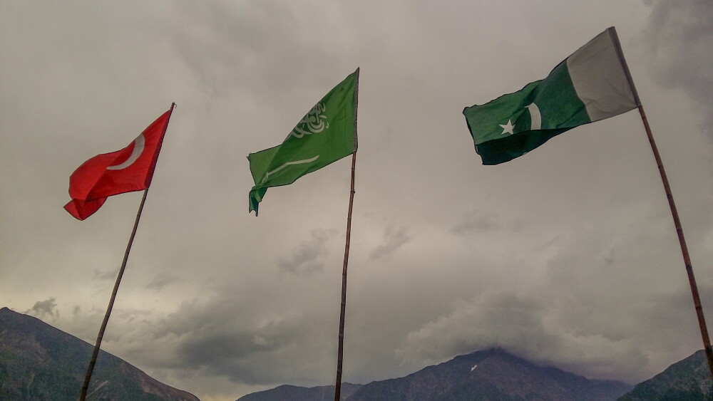 The national flags of Turkey, Saudi Arabia, and Pakistan flutter against a backdrop of cloudy skies and mountainous terrain.