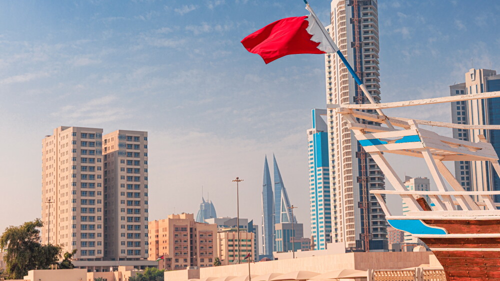 A flag of Bahrain flutters on a traditional dhow, as Manama's modern buildings create a dynamic cityscape backdrop.