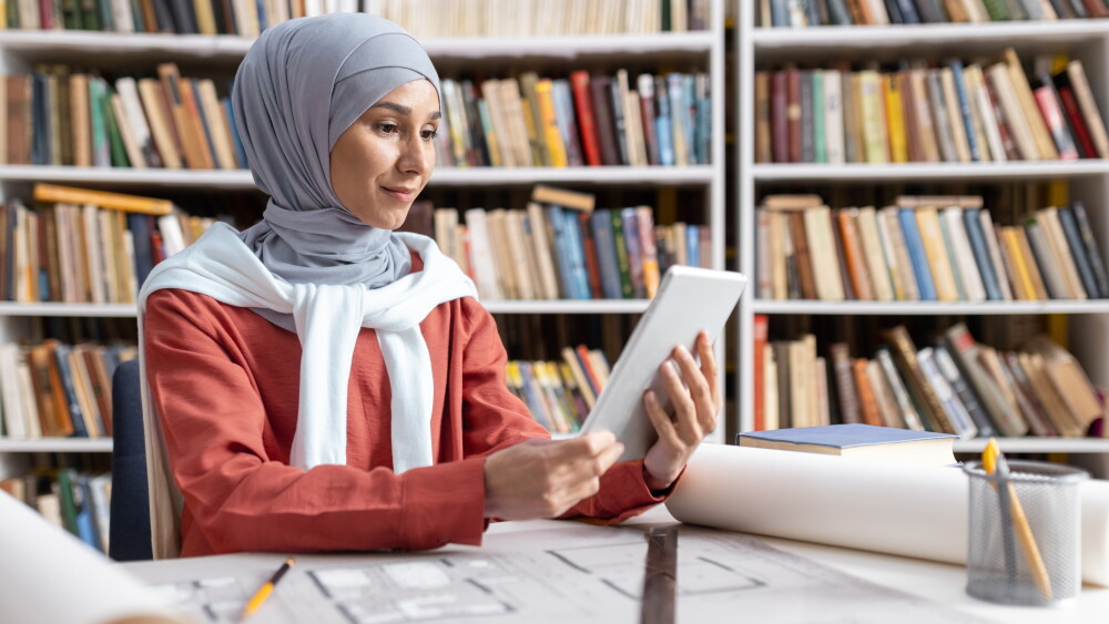 A young Muslim woman consults a tablet in a library.