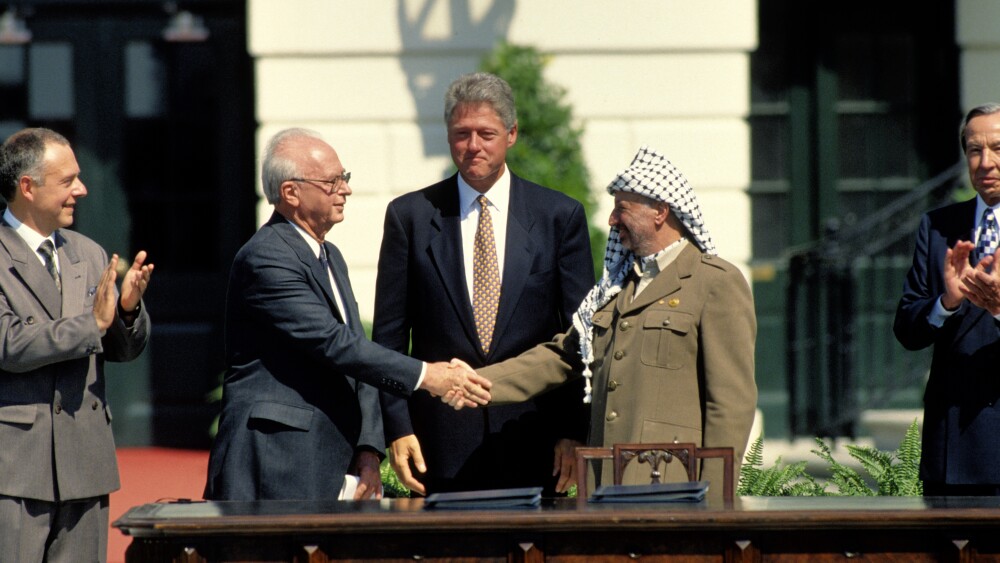 On September 13, 1993, Yitzhak Rabin of Israel and Yasser Arafat of the PLO shake hands at the White House South Lawn after signing the Oslo Peace Accords; President Bill Clinton looks on.