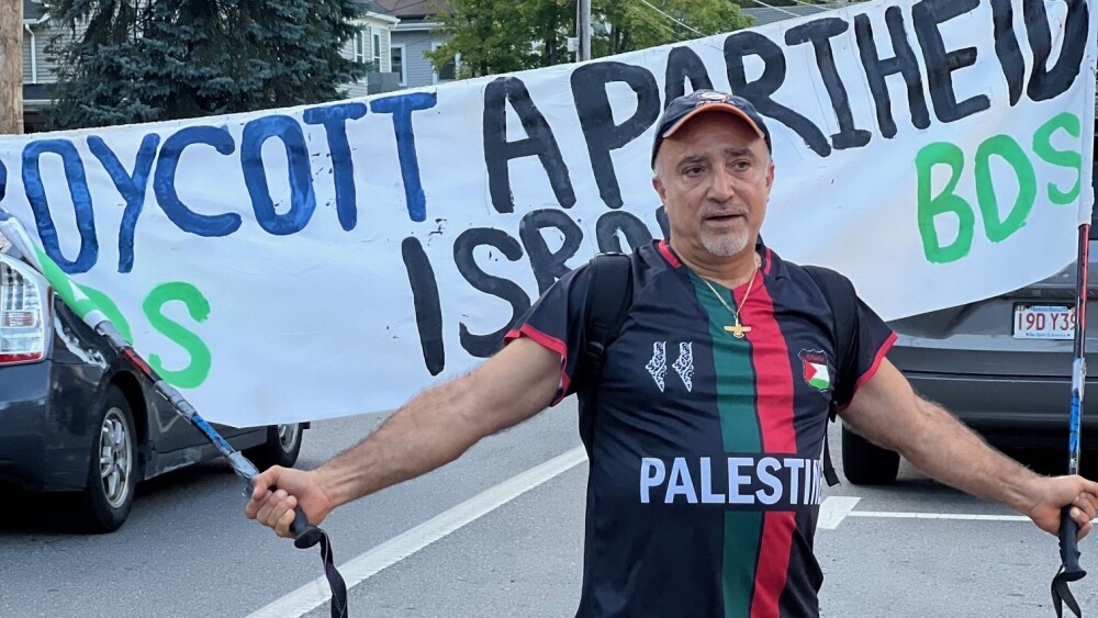 Kian Kianfar, an Iranian-born American who lives in Framingham, Mass., appears to relish his status as a mascot for the oppressive Iranian regime at protests in the suburbs of Boston. Above, he carries a banner accusing Israel of apartheid while taunting his adversaries at a rally in Lexington, Mass., in mid-August 2024.