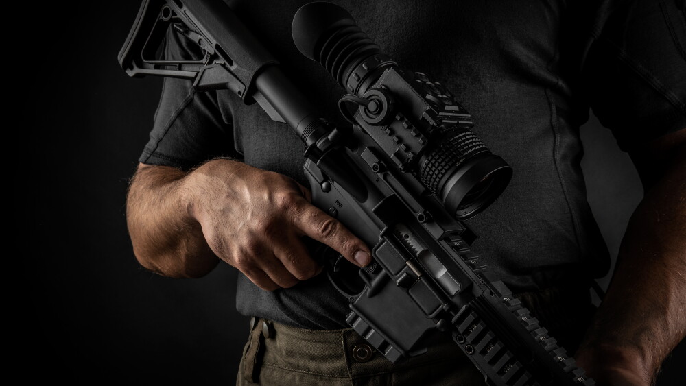 A man holds an automatic carbine with an optical sight and a night vision device.